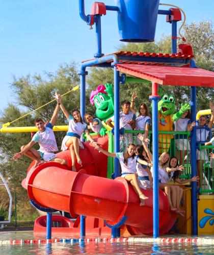Gruppo felice in parco acquatico con scivoli colorati e mascotte verde.